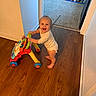 baby, walker, toy, wooden_floor, carpet, bed, superhero_bed, smile, standing, indoor, child, room, happy, footwear, pajamas, furniture, wall, doorway, playroom, infant