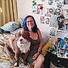 Banksy is registered to the contest to win money with this photo: art_supplies, artwork, bed, bedroom, blanket, blue_hair, brown_dog, cozy, dog, glasses, indoors, person, pet, pillows, portrait, smiling, tattoo, wall_decor, white_dog, woman