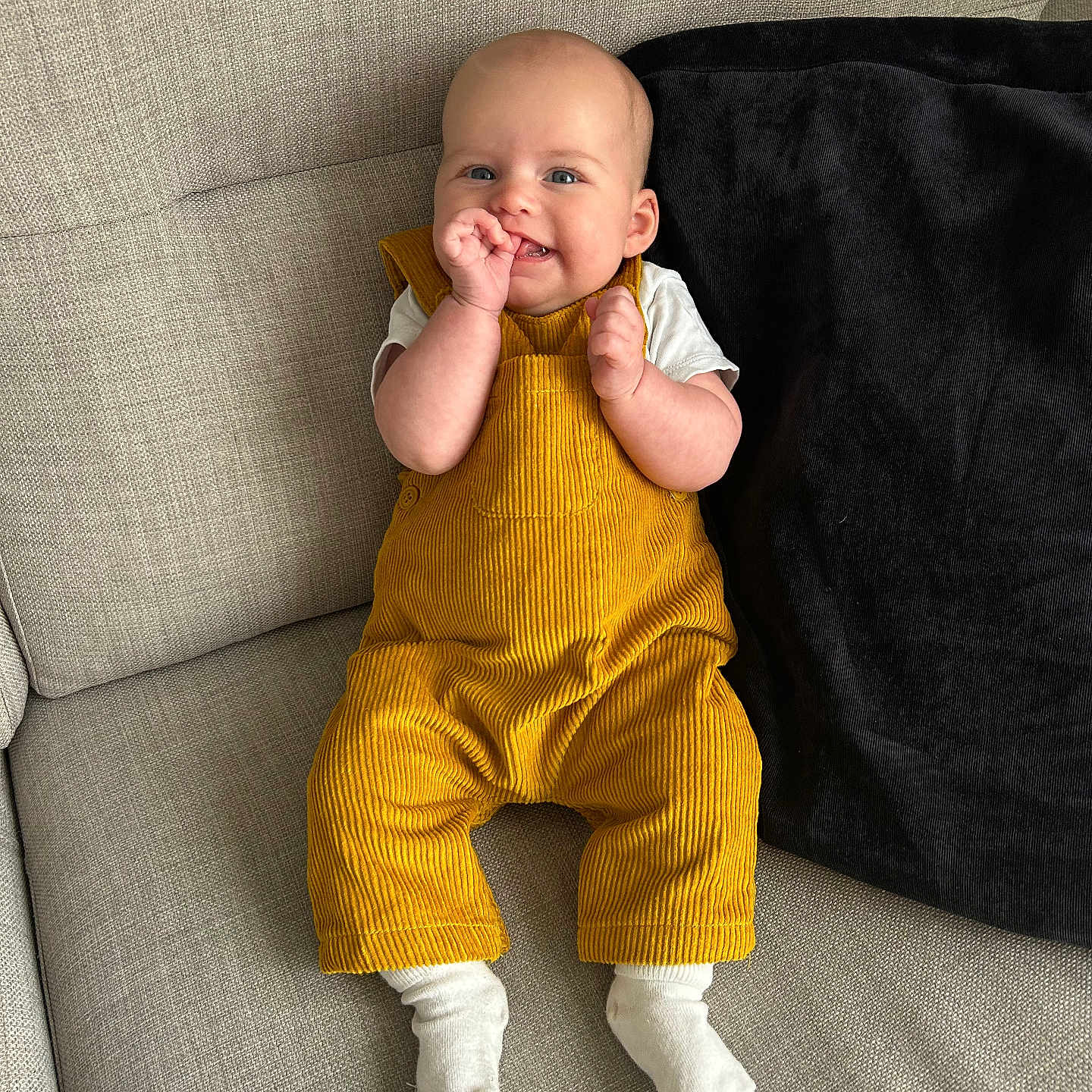 Myla joined the competition — help win amazing prizes! baby, child, clothing, comfortable, couch, cute, furniture, happy, home, indoor, infant, person, pillow, playful, portrait, seated, smiling, white_socks, yellow_overalls, young_child