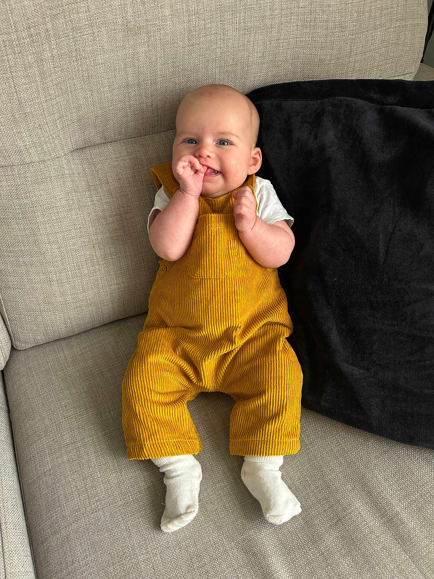 Myla joined the competition — help win amazing prizes! baby, child, smiling, yellow_overalls, white_socks, couch, pillow, indoor, portrait, happy, infant, clothing, person, seated, cute, playful, furniture, home, comfortable, young_child