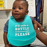 Amir joined the competition — help win amazing prizes! baby, child, bib, smiling, indoor, person, hand, bracelet, curly_hair, clothing, portrait, standing, face, expression, background, sign, flag, support, cute, infant