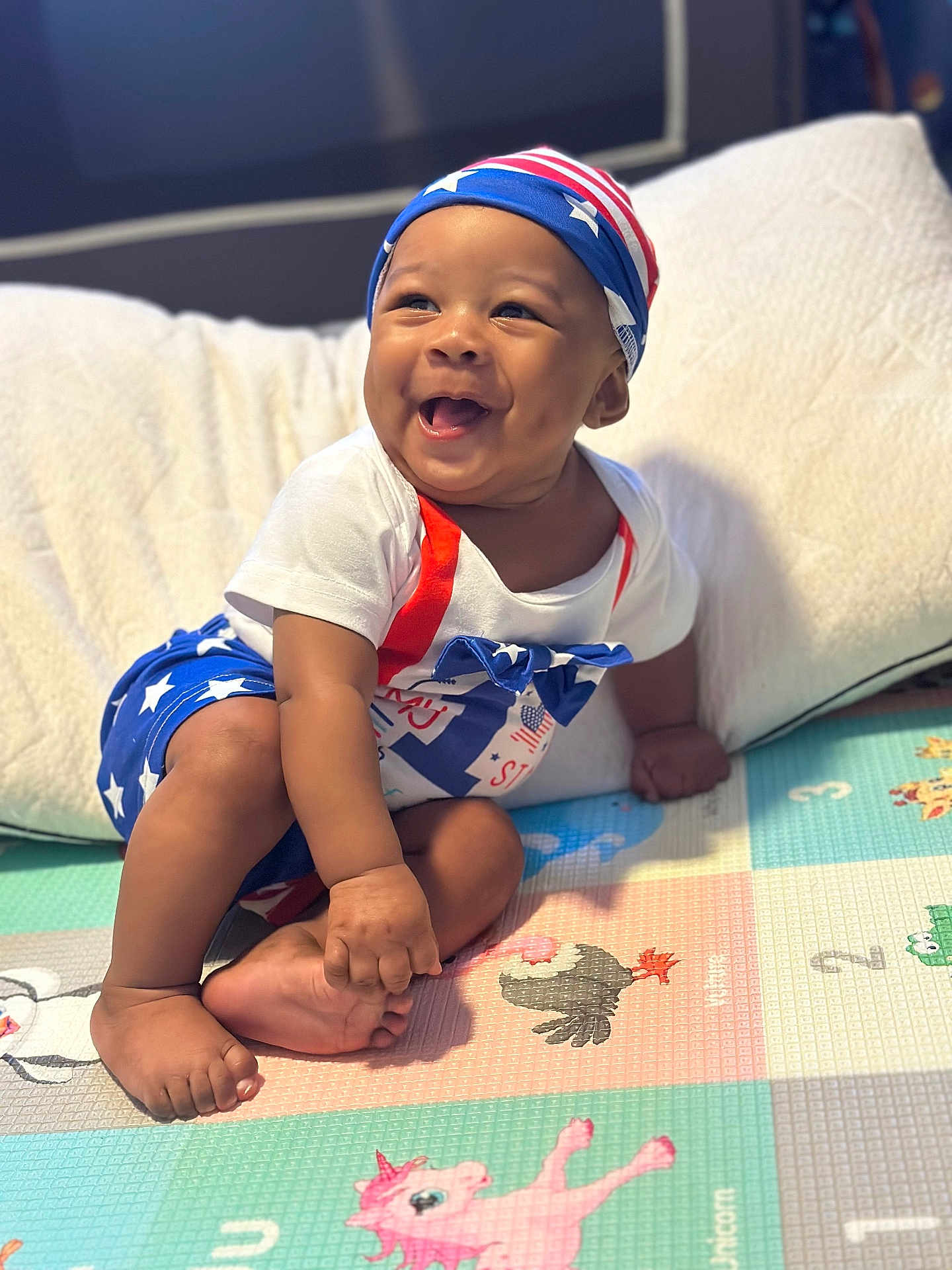 Nehemiah joined the competition — help win amazing prizes! baby, child, smiling, sitting, play_mat, colorful, patriotic_clothing, stars, stripes, headwear, happy, indoors, foot, hand, person, infant, cushion, clothing, cute, portrait