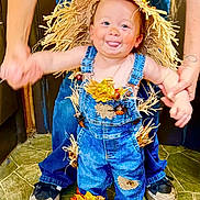 Ripp is registered to the contest to win money with this photo: toddler, child, scarecrow_costume, denim_overalls, straw, autumn_leaves, playful, tongue_out, hands_held, floor, indoor, person, cute, smiling, hat, footwear, jeans, holding_hands, happy, support