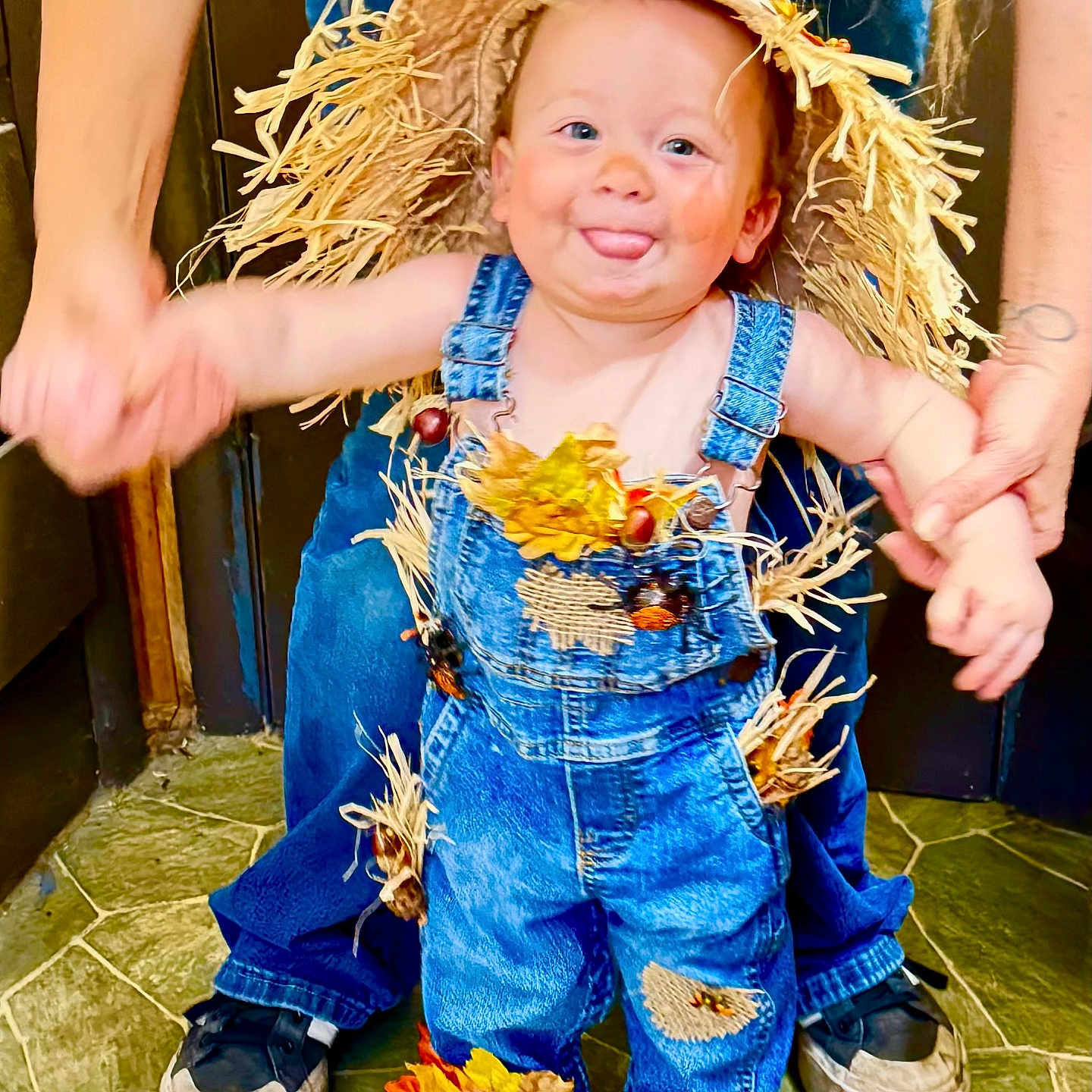Ripp is registered to the contest to win money with this photo: autumn_leaves, child, cute, denim_overalls, floor, footwear, hands_held, happy, hat, holding_hands, indoor, jeans, person, playful, scarecrow_costume, smiling, straw, support, toddler, tongue_out