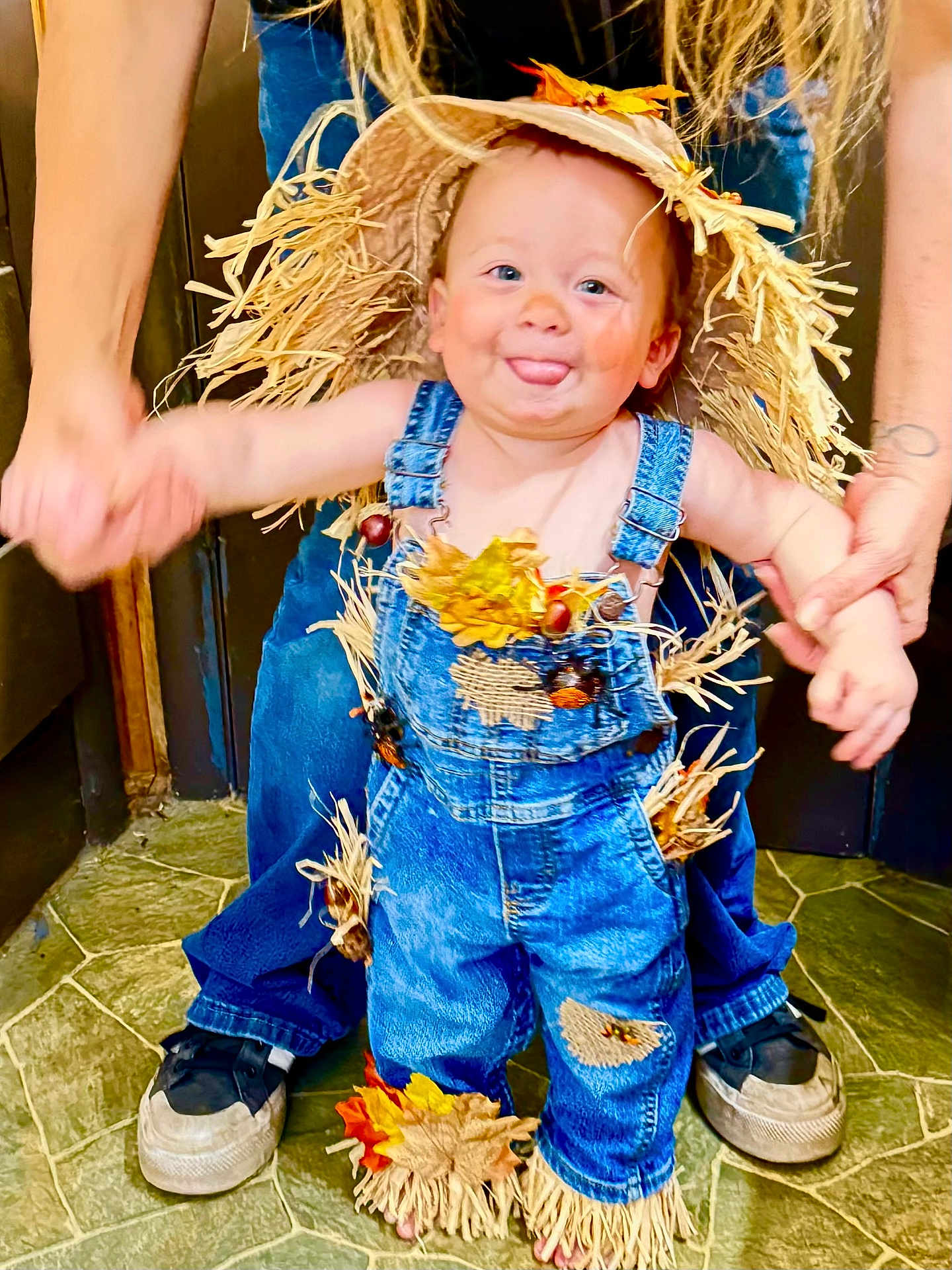 Ripp is registered to the contest to win money with this photo: toddler, child, scarecrow_costume, denim_overalls, straw, autumn_leaves, playful, tongue_out, hands_held, floor, indoor, person, cute, smiling, hat, footwear, jeans, holding_hands, happy, support