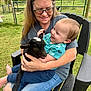 toddler, child, woman, glasses, baby_goat, animal, outdoor, grass, fence, black_chair, smiling, holding, happy, casual_clothing, nature, daylight, playing, affection, portrait, people