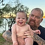 baby, smiling, man, tattoos, beard, outdoor, lake, water, trees, nature, sunset, holding, happy, child, portrait, skin, person, grass, father, family