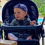 baby, boy, stroller, cap, outdoor, greenery, pool, child, infant, seat, summer, casual_clothing, expression, curious, sunlight, nature, daytime, portrait, young_child, relaxing