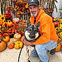 Maxx is registered to the contest to win money with this photo: man, dog, pumpkins, flowers, cornstalks, orange_hoodie, leash, indoor, fall, autumn, smiling, beard, cap, jeans, boots, decor, hay, black_and_white_dog, crouching, pet