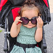 Sophia is registered to the contest to win money with this photo: toddler, child, sunglasses, stroller, green_dress, outdoor, cute, smiling, baby, fashion, seatbelt, hair_bun, playful, summer, portrait, person, seat, daylight, fun, accessory