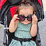 toddler, child, sunglasses, stroller, green_dress, outdoor, cute, smiling, baby, fashion, seatbelt, hair_bun, playful, summer, portrait, person, seat, daylight, fun, accessory