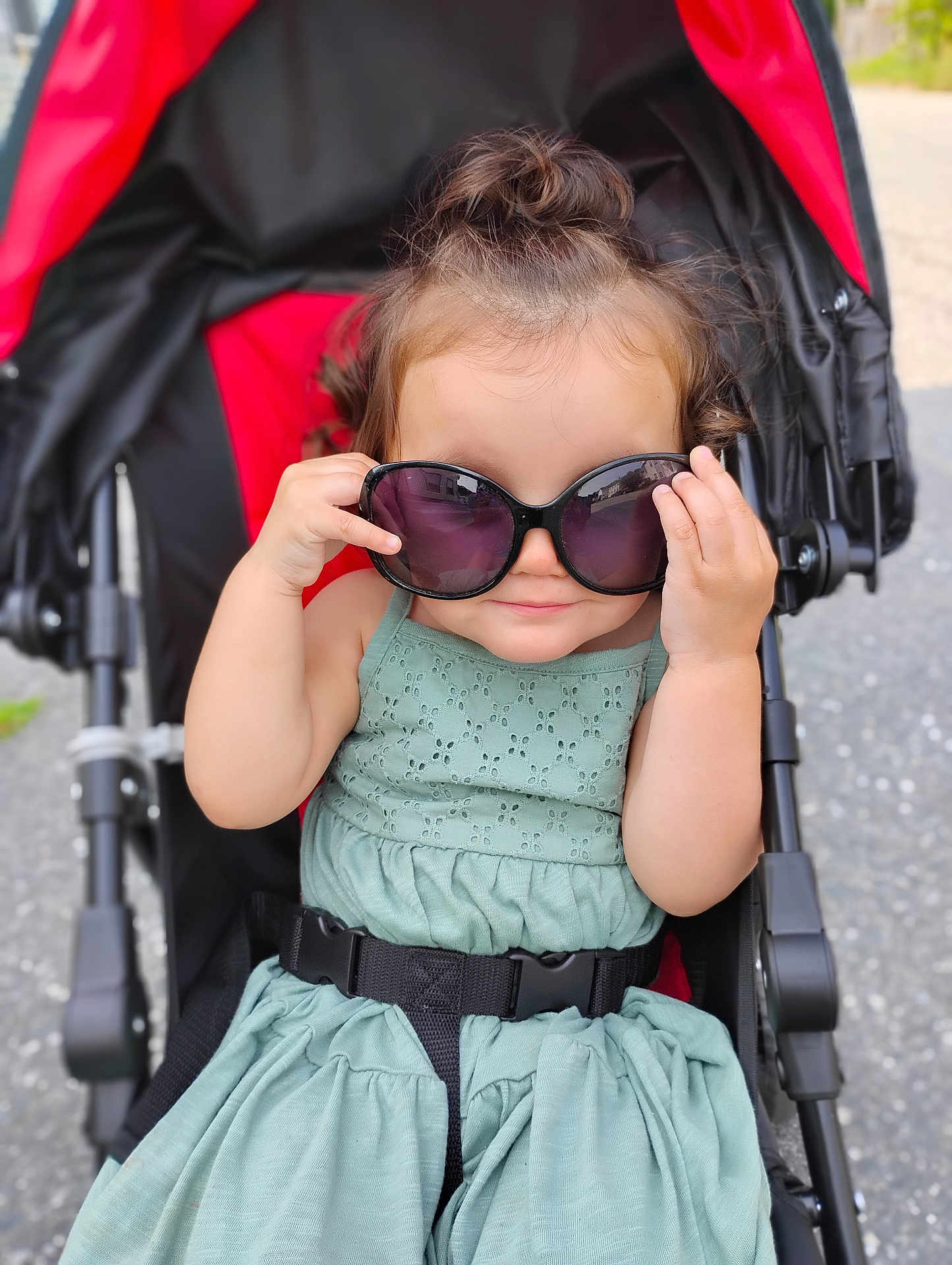 Sophia is registered to the contest to win money with this photo: toddler, child, sunglasses, stroller, green_dress, outdoor, cute, smiling, baby, fashion, seatbelt, hair_bun, playful, summer, portrait, person, seat, daylight, fun, accessory