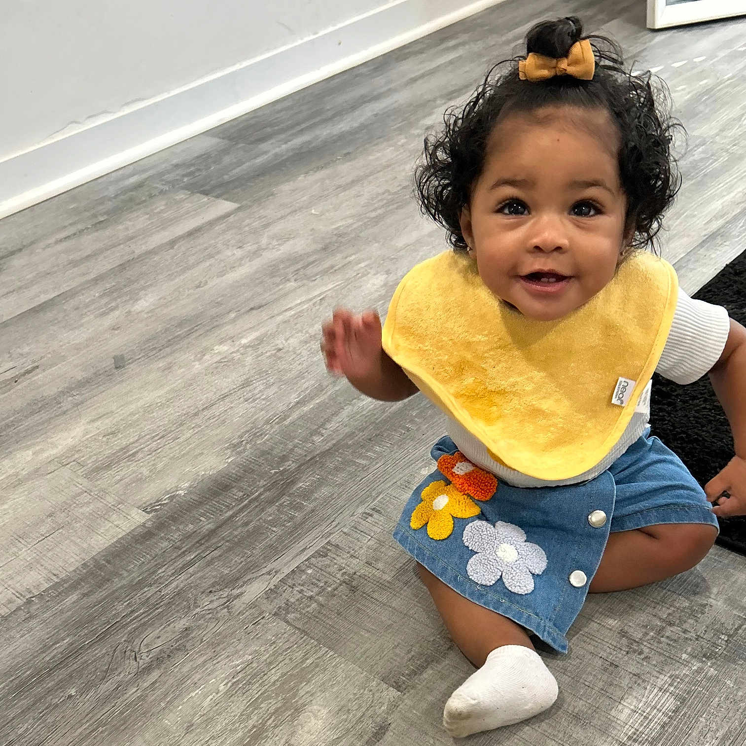 Lylann is registered to the contest to win money with this photo: baby, black_rug, child, curly_hair, cute, denim_skirt, floor, flooring, flower_patch, hair_bow, happy, indoor, person, portrait, sitting, smiling, sock, toddler, white_shirt, yellow_bib