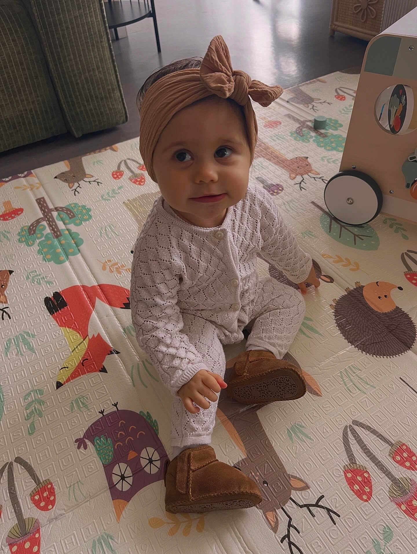 Anaë a rejoint le concours — aidez-le/la à gagner de superbes lots ! child, baby, toddler, headband, bow, onesie, knitted_clothing, moccasins, playmat, sitting, indoor, toy_cart, cute_expression, big_eyes, floor, patterned_mat, fox_illustration, hedgehog_illustration, owl_illustration, soft_lighting
