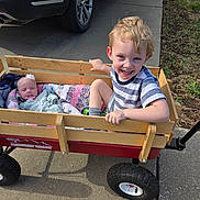 Sammy is registered to the contest to win money with this photo: child, baby, wagon, wooden_wagon, smiling, sleeping, blanket, headband, flower, outdoor, sidewalk, car, toddler, boy, infant, happy, person, feet, shoe, sunlight