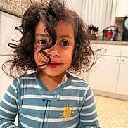 Valentina is registered to the contest to win money with this photo: child, toddler, curly_hair, striped_clothing, blue_onesie, sun_patch, kitchen, countertop, appliances, indoor, portrait, smile, hands, cute, young, person, face, curious, home, lighting