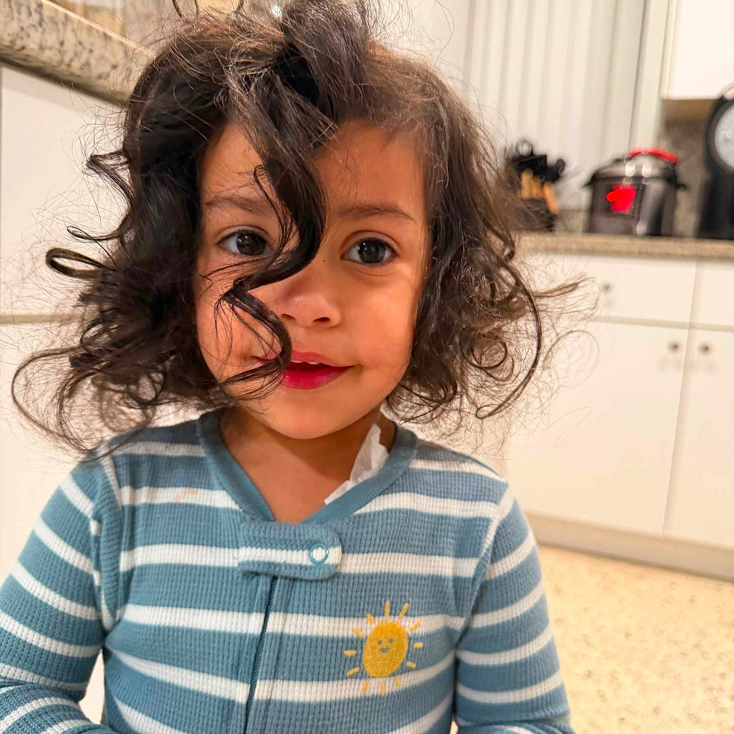 Valentina is registered to the contest to win money with this photo: child, toddler, curly_hair, striped_clothing, blue_onesie, sun_patch, kitchen, countertop, appliances, indoor, portrait, smile, hands, cute, young, person, face, curious, home, lighting