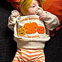 baby, infant, pacifier, headband, yellow_headband, pumpkin, pumpkin_design, sweater, striped_pants, orange_stripes, cute, lying_down, pillow, orange_pillow, cozy, child, indoors, soft_lighting, smiling_pumpkins, clothing
