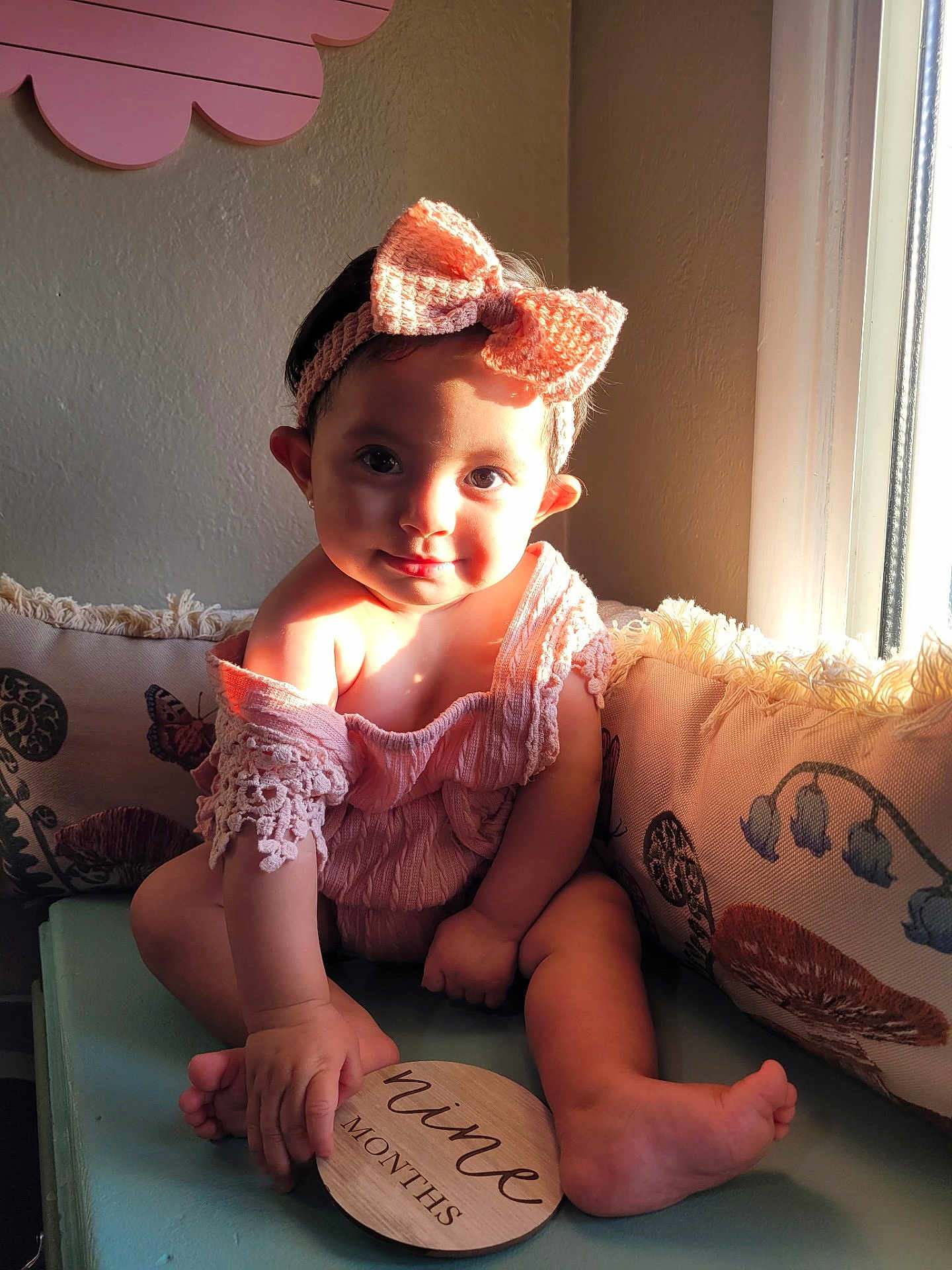 Frannie joined the competition — help win amazing prizes! baby, child, girl, headband, bow, pink_outfit, cushion, bench, window, sunlight, wooden_sign, nine_months, flower_wall_decor, indoor, cute, smiling, sitting, hand, foot, portrait