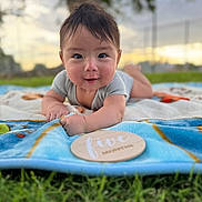 Owen is registered to the contest to win money with this photo: baby, infant, smiling, blanket, outdoor, grass, wooden_plaque, milestone, face, portrait, child, nature, sunset, cute, happy, toddler, young_child, clothing, head, expression