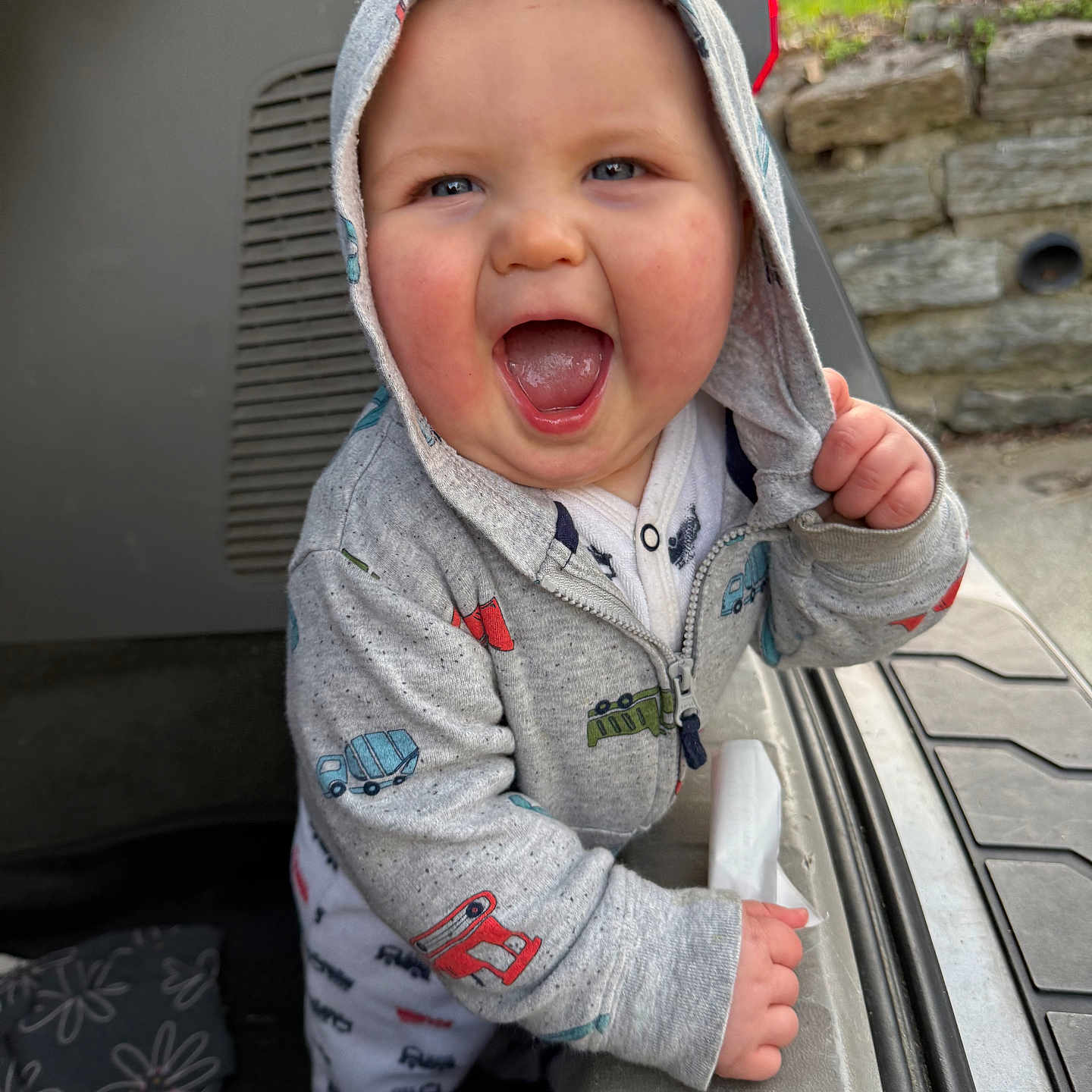 Colter is registered to the contest to win money with this photo: baby, child, hoodie, grey_clothing, smiling, open_mouth, blue_eyes, hand, car_trunk, outdoor, daylight, stone_wall, greenery, grass, playful, happy, infant, cute, person, casual_wear