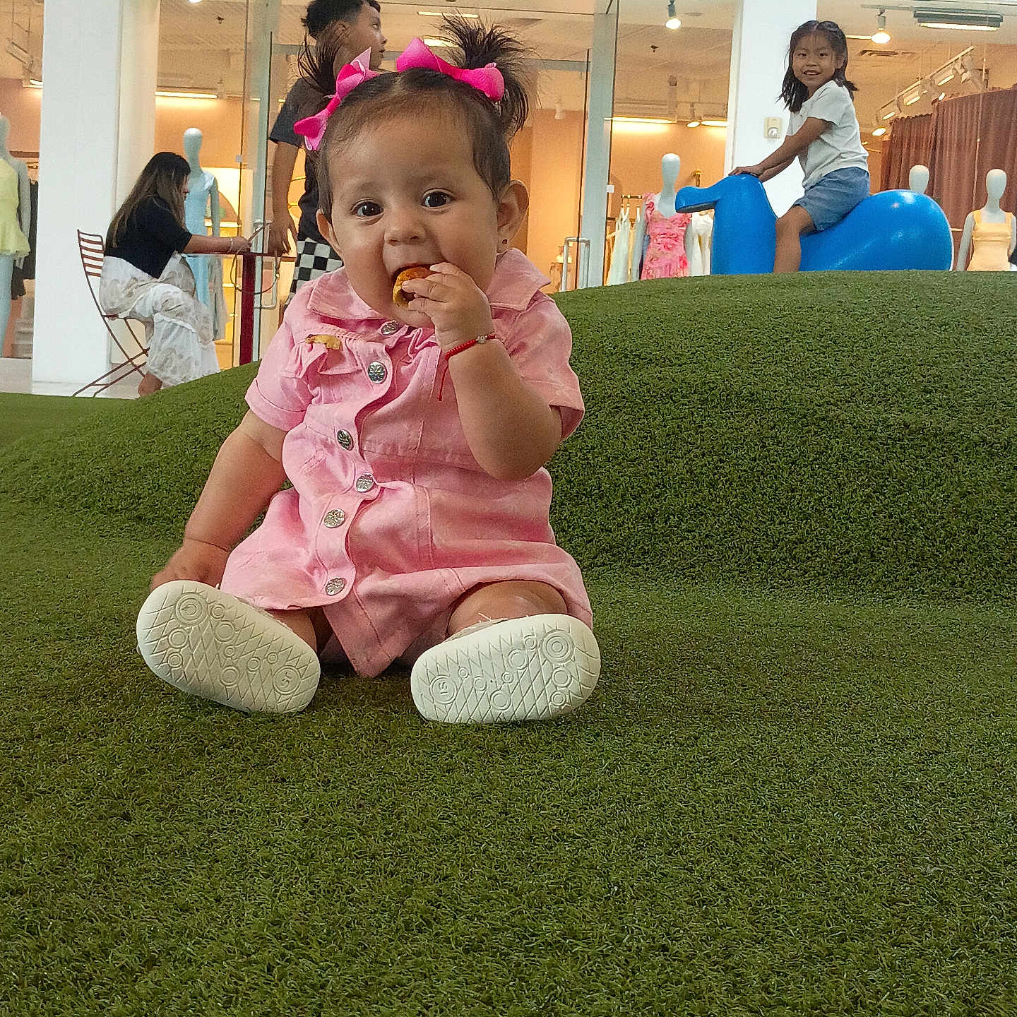 Maria Catalina is registered to the contest to win money with this photo: baby, child, toddler, girl, pink_dress, hair_bows, snack, grass, indoor, play_area, toy_horse, smiling, shop_window, mannequins, people, seated, happy, casual_clothing, retail_store, green