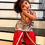 child, curly_hair, traditional_clothing, red_pants, patterned_top, barefoot, stairs, indoors, pose, cute, portrait, young_child, smiling, expression, playful, home, person, fashion, colorful, happy