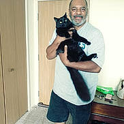 Carl E. is registered to the contest to win money with this photo: animal, black_cat, carpet, domestic, door, furniture, happy, holding, home, indoor, man, pet, room, shoes, shorts, smile, standing, table, tail, tshirt