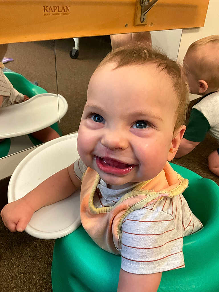 Beau is registered to the contest to win money with this photo: baby, child, smiling, blue_eyes, bib, green_seat, mirror, reflection, crawling, carpet, indoor, toddler, happy, face, person, short_sleeve_shirt, striped_shirt, infant, cute, playroom