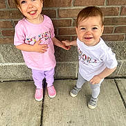 Piper is registered to the contest to win money with this photo: toddler, child, holding_hands, smiling, outdoor, sidewalk, brick_wall, concrete, pink_shirt, white_shirt, purple_pants, gray_pants, shoes, cute, happy, person, portrait, two_children, young_child, casual_clothing