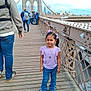 Ravisha is registered to the contest to win money with this photo: child, girl, bridge, brooklyn_bridge, walking, people, jeans, purple, hair_bows, sidewalk, outdoor, cityscape, sky, daytime, casual_clothing, smile, necklace, urban, tourist, pedestrian