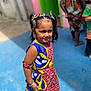 child, girl, colorful_dress, headband, standing, outdoor, blue_floor, people, background_blur, smiling, portrait, sunlight, patterned_clothing, hair_tied, accessories, happy, casual, vibrant_colors, person, fashion