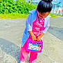 child, kid, toy, medical_kit, doctor_costume, outdoor, sidewalk, shoes, pink_clothing, white_coat, hair_bun, sunny_day, greenery, play, pretend_play, young_child, standing, casual, bright_colors, street