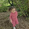 child, toddler, pink_jumpsuit, outdoor, garden, trees, greenery, wood_chips, nature, curious, walking, daylight, plants, young_child, casual_clothing, playful, hair_tied, leaves, bushes, exploring