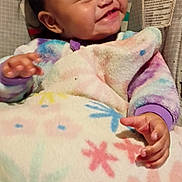 Billie is registered to the contest to win money with this photo: baby, baby_seat, blanket, child, clothing, colorful, cozy, cute, expression, face, floral_pattern, hand, happy, indoor, infant, monkey_illustration, person, portrait, smiling, young_child