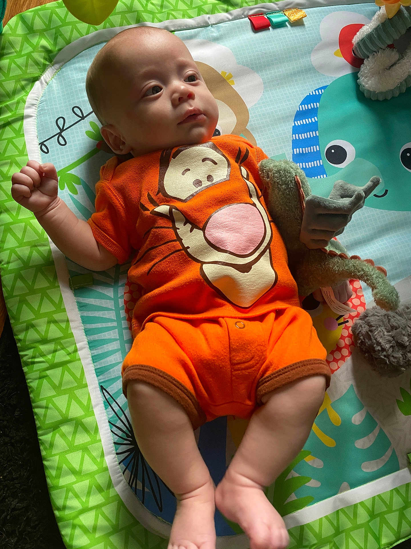 Elijah joined the competition — help win amazing prizes! baby, infant, onesie, orange_clothing, plush_toy, play_mat, tigger, laying_down, child, cute, soft_light, indoor, colorful, expression, person, legs, arms, face, toy, floor_mat