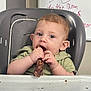 Nashton is registered to the contest to win money with this photo: baby, child, high_chair, meat, food, eating, face, hands, messy, indoor, whiteboard, writing, curious, young_child, green_shirt, table, person, light, skin, expression