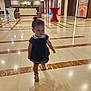 toddler, child, walking, marble_floor, lobby, indoor, dress, sandals, shadow, artwork, table, plant, ceiling_lights, person, hallway, wall_art, furniture, young_child, small_person, interior