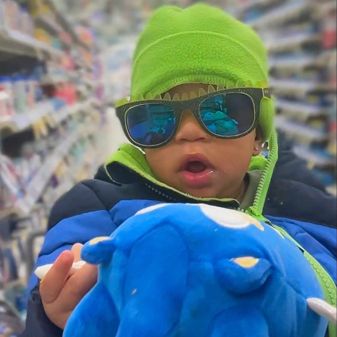 Truu Daniels is registered to the contest to win money with this photo: accessories, aisle, baby, blue_plush, child, close_up, clothing, earring, green_beanie, grocery_store, hat, plush_toy, portrait, reflection_in_glasses, shopping_cart, stuffed_animal, sunglasses, surprised_expression, toddler, winter_coat