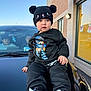 toddler, child, black_clothing, hat, shoes, car, hood, building, window, people, outdoor, daylight, brick_wall, sneakers, face, person, casual, urban, sky, portrait
