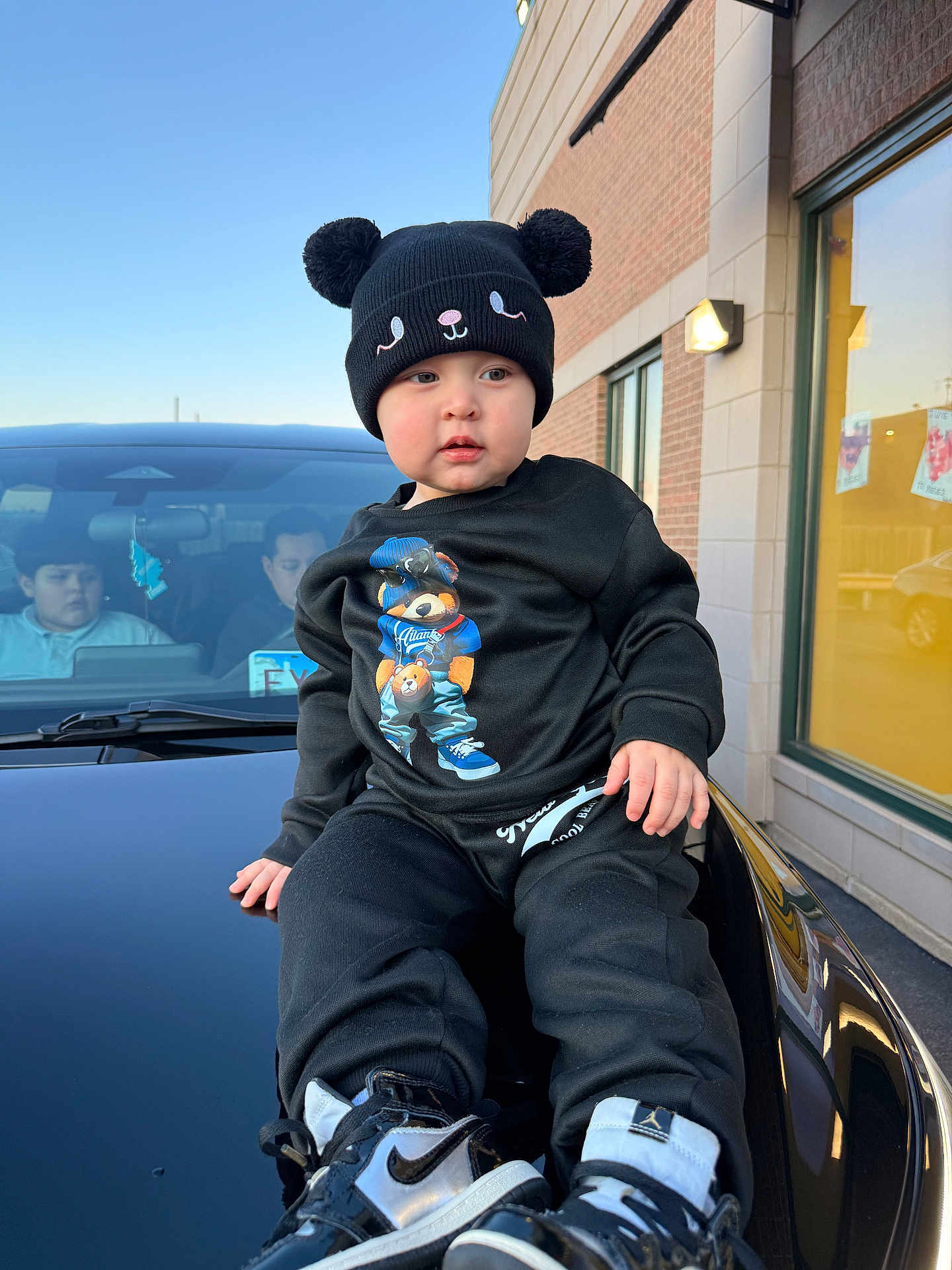 Thiago is registered to the contest to win money with this photo: toddler, child, black_clothing, hat, shoes, car, hood, building, window, people, outdoor, daylight, brick_wall, sneakers, face, person, casual, urban, sky, portrait