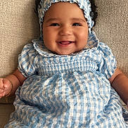 Juliana is registered to the contest to win money with this photo: baby, child, smiling, curly_hair, headband, blue_dress, checkered_pattern, sitting, couch, texture, happy, infant, portrait, indoors, cute, fashion, young, person, skin, face