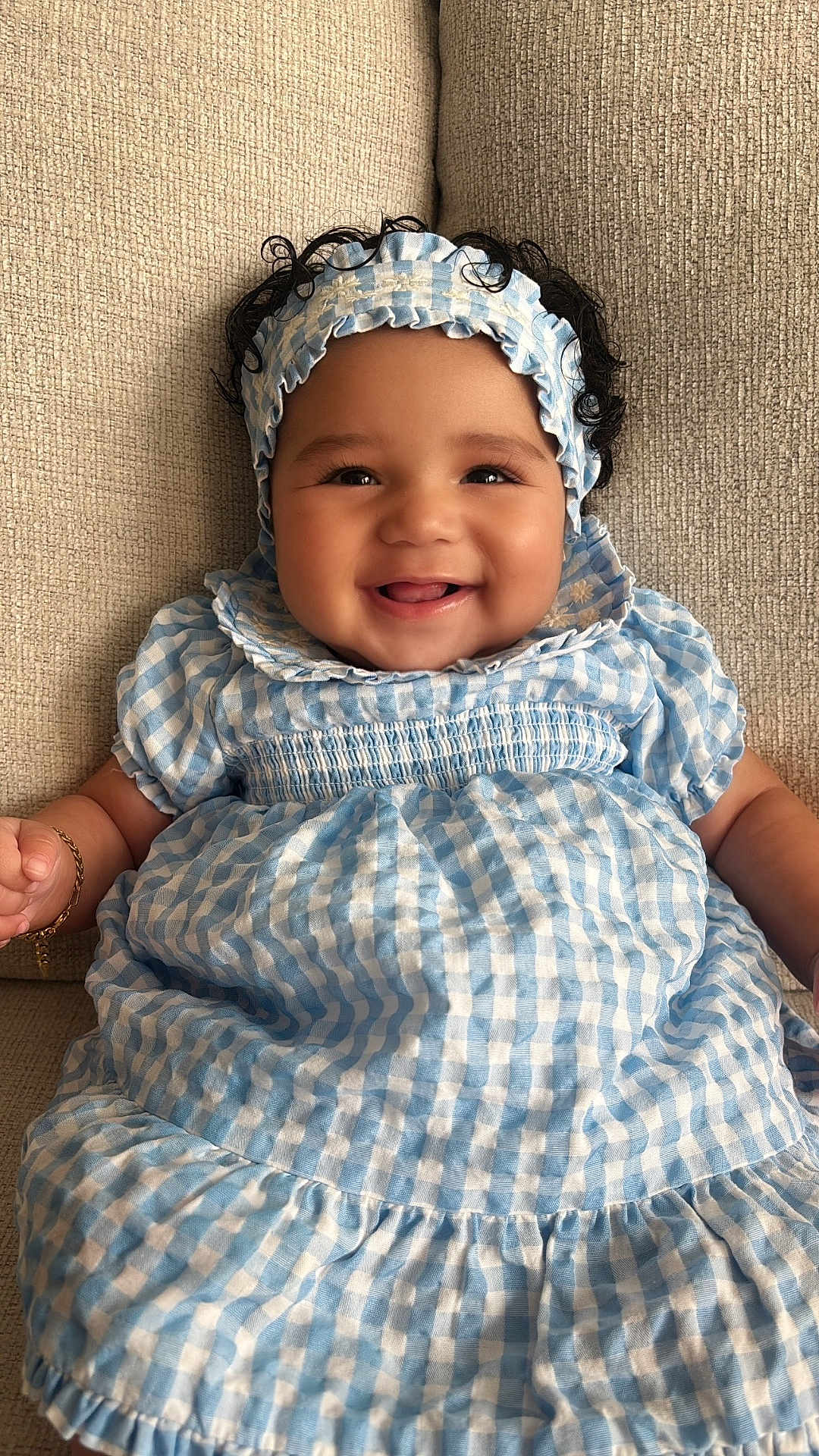Juliana is registered to the contest to win money with this photo: baby, child, smiling, curly_hair, headband, blue_dress, checkered_pattern, sitting, couch, texture, happy, infant, portrait, indoors, cute, fashion, young, person, skin, face