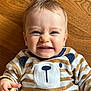 baby, child, face, smile, expression, striped_clothing, bear_design, wooden_floor, happy, cute, infant, portrait, playful, sitting, person, clothing, baby_clothes, indoors, skin, hand