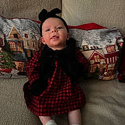 Lynette is registered to the contest to win money with this photo: baby, infant, child, couch, pillow, plaid_dress, black_headband, mittens, white_socks, holiday_theme, christmas_pillow, indoor, soft_lighting, portrait, smiling, sitting, cozy, cute, person, home