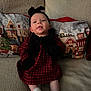 baby, infant, child, couch, pillow, plaid_dress, black_headband, mittens, white_socks, holiday_theme, christmas_pillow, indoor, soft_lighting, portrait, smiling, sitting, cozy, cute, person, home