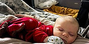 Lynette is registered to the contest to win money with this photo: baby, sleeping, pajamas, red_clothing, blanket, cushion, bed, indoor, cozy, child, peaceful, infant, soft_texture, warm_lighting, face, hand, rest, cute, nursery, comfort