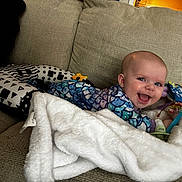 Lynette is registered to the contest to win money with this photo: baby, infant, smile, tummy_time, couch, blanket, toy, playmat, pillow, living_room, happy, cute, face, onesie, patterned_clothing, soft_texture, home_interior, pacifier, cheerful, photography