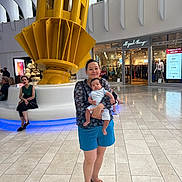 David joined the competition — help win amazing prizes! architecture, baby, background, casual_clothing, clothing_store, floor, footwear, holding, indoor, light, modern, people, portrait, public_space, shopper, shopping_mall, storefront, tiles, woman, yellow_structure