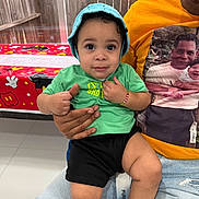 Andy is registered to the contest to win money with this photo: toddler, child, baby, hat, green_tshirt, black_shorts, bracelet, adult, orange_tshirt, printed_photo, mickey_mouse, tablecloth, indoor, person, sitting, jeans_shorts, hand, face, floor, window